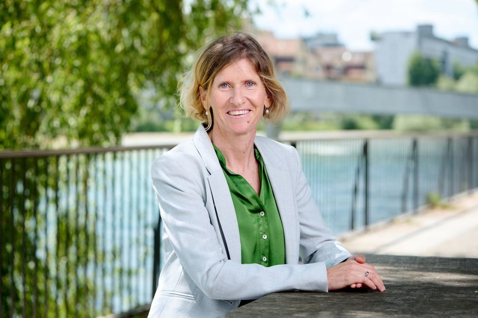 Die Frau posiert an einem Flussufer und trägt eine grüne Bluse mit einem hellgrauen Blazer.